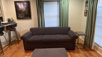 A living room with a sofa, a small table, a bar stool, a window with curtains, and a framed picture on the wall.