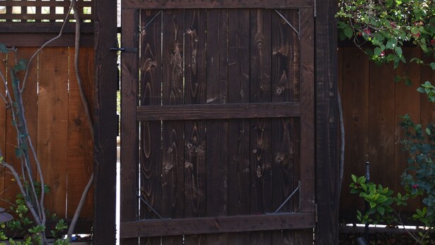 Una reja de madera con un arco de madera encima, rodeada de abundante follaje y plantas con flores.