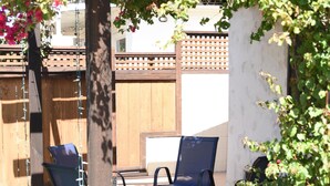 Un espacio al aire libre con un porche de madera cubierto de enredaderas florecientes, una zona de estar con sillas azules y una cerca de madera al fondo.