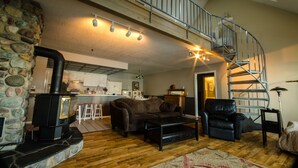 A cozy living room with a fireplace, a staircase, and a kitchen in the background. - Lakefront Home With Hot Tub, Dock, Great Views From The Living Room & Bedrooms  (Somers)