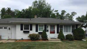 A single-story house with a red front door, white siding, and black shutters. There are two small evergreen trees in front of the house.