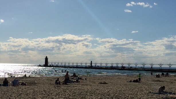 A beach with people relaxing, a pier extending into the water, and a lighthouse in the background.