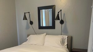 A neatly made bed with white linens, two wall-mounted reading lamps, and a framed mirror above it. - Downtown/Corporate/Individuals (Anchorage)