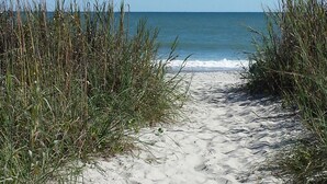 A sandy beach path leading to the ocean, surrounded by tall grass.