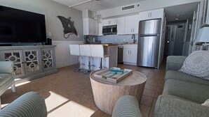 A modern living room with a flat-screen TV, white furniture, a kitchen area with stainless steel appliances, and a dining area with bar stools.