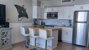A modern kitchen with white cabinets, a stainless steel refrigerator, a microwave, and a large flat-screen TV. There are two bar stools and a wooden countertop.