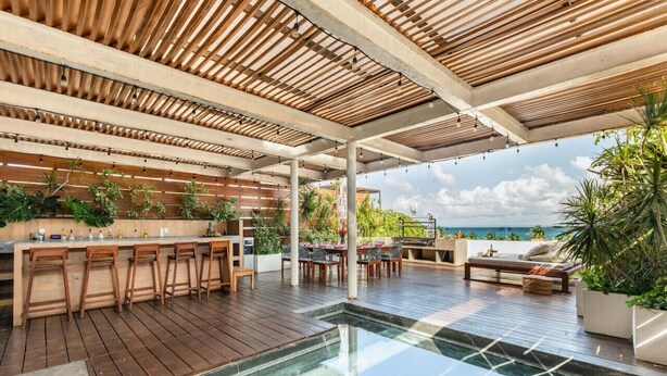 Un área de piscina al aire libre con cubierta de madera, un bar con taburetes y una zona de estar con mesa y sillas.