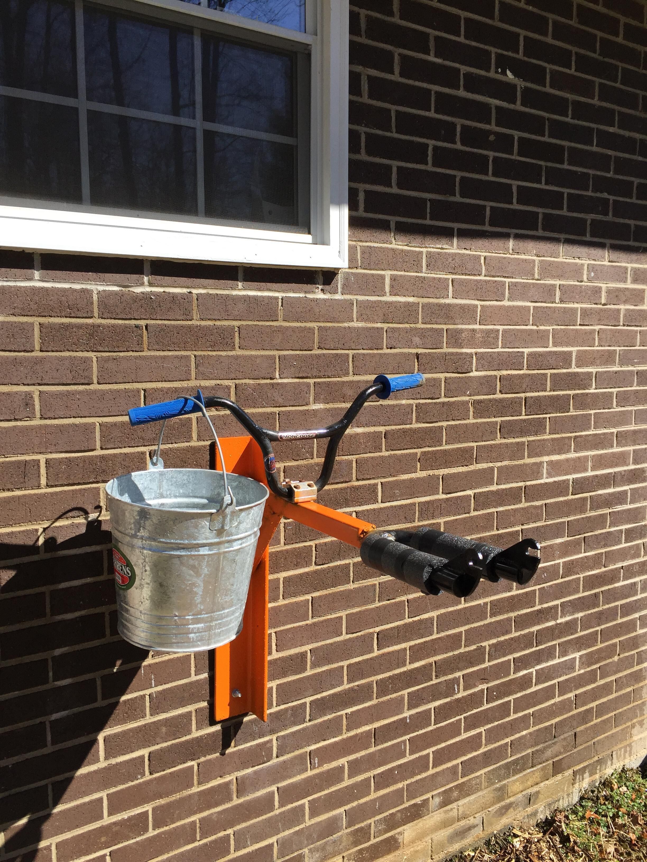 A wheelbarrow with a metal bucket attached to it, leaning against a brick wall with a window above.