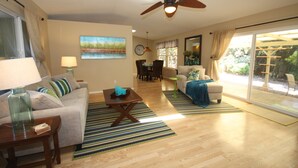 A spacious living room with a ceiling fan, a dining area, and a sliding glass door leading to a patio. - Magic Sands Home, Steps From The Beach! Air Conditioned! 3D tour to view space (Kailua-Kona)