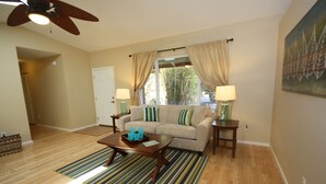 A living room with a beige sofa, a wooden coffee table, and a striped rug. There is a ceiling fan, a window with curtains, and a painting on the wall. - Magic Sands Home, Steps From The Beach! Air Conditioned! 3D tour to view space (Kailua-Kona)