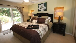 A bedroom with a bed, a ceiling fan, a nightstand with a lamp, and a window with curtains. - Magic Sands Home, Steps From The Beach! Air Conditioned! 3D tour to view space (Kailua-Kona)