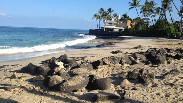 A beach with large rocks in the foreground, a building with a balcony and palm trees in the background, and the ocean waves. - Magic Sands Home, Steps From The Beach! Air Conditioned! 3D tour to view space (Kailua-Kona)