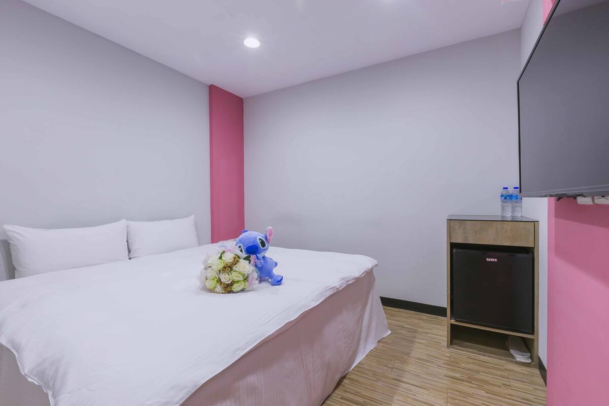 Una habitació d'hotel amb un llit, una petita taula de treball amb una televisió, una paret rosa i un sòl de fusta.