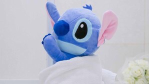 Un peluche de Stitch envuelto en una toalla blanca sobre un suelo de baldosas.