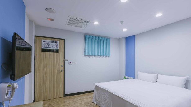 A hotel room with a bed, a wooden door, a blue curtain, a wall-mounted TV, and a hairdryer on the wall. - Mei Ti Hotel (Taoyuan City)