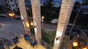 Un'area salotto esterna con mobili in vimini e pavimento piastrellato, circondata da colonne in pietra e illuminata da lampade a sospensione.