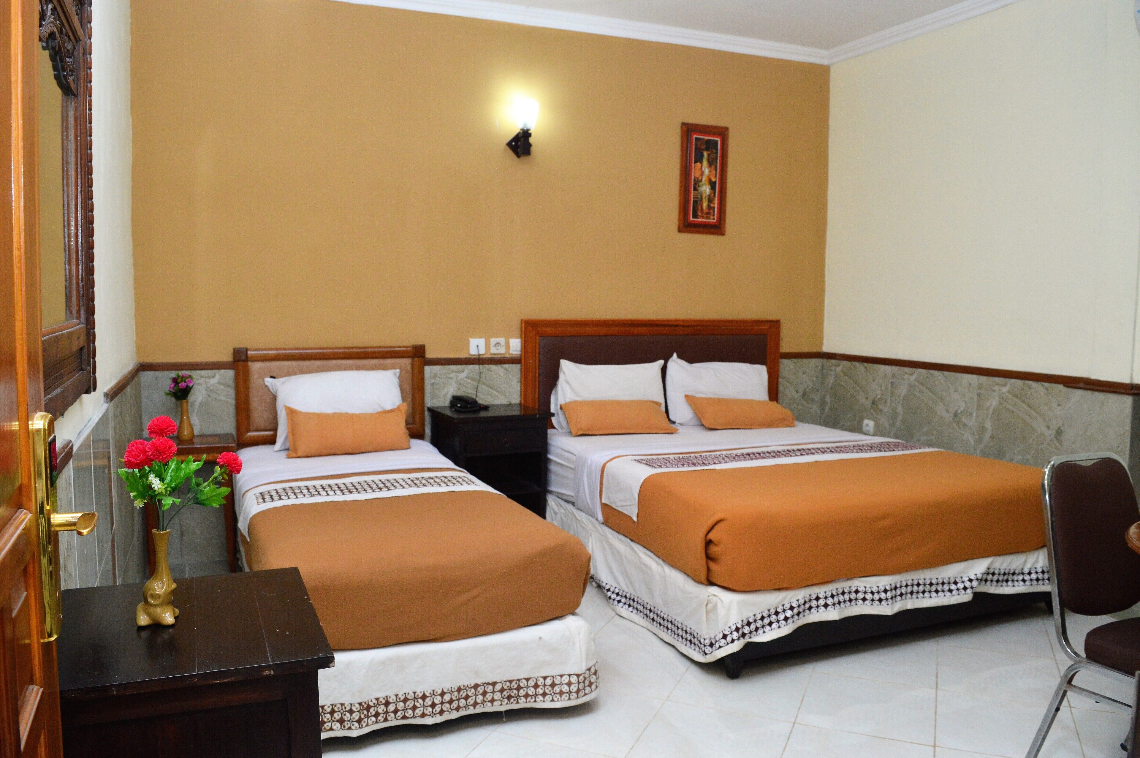 A hotel room with two beds, a nightstand, a chair, a vase with flowers, and a wall-mounted light.