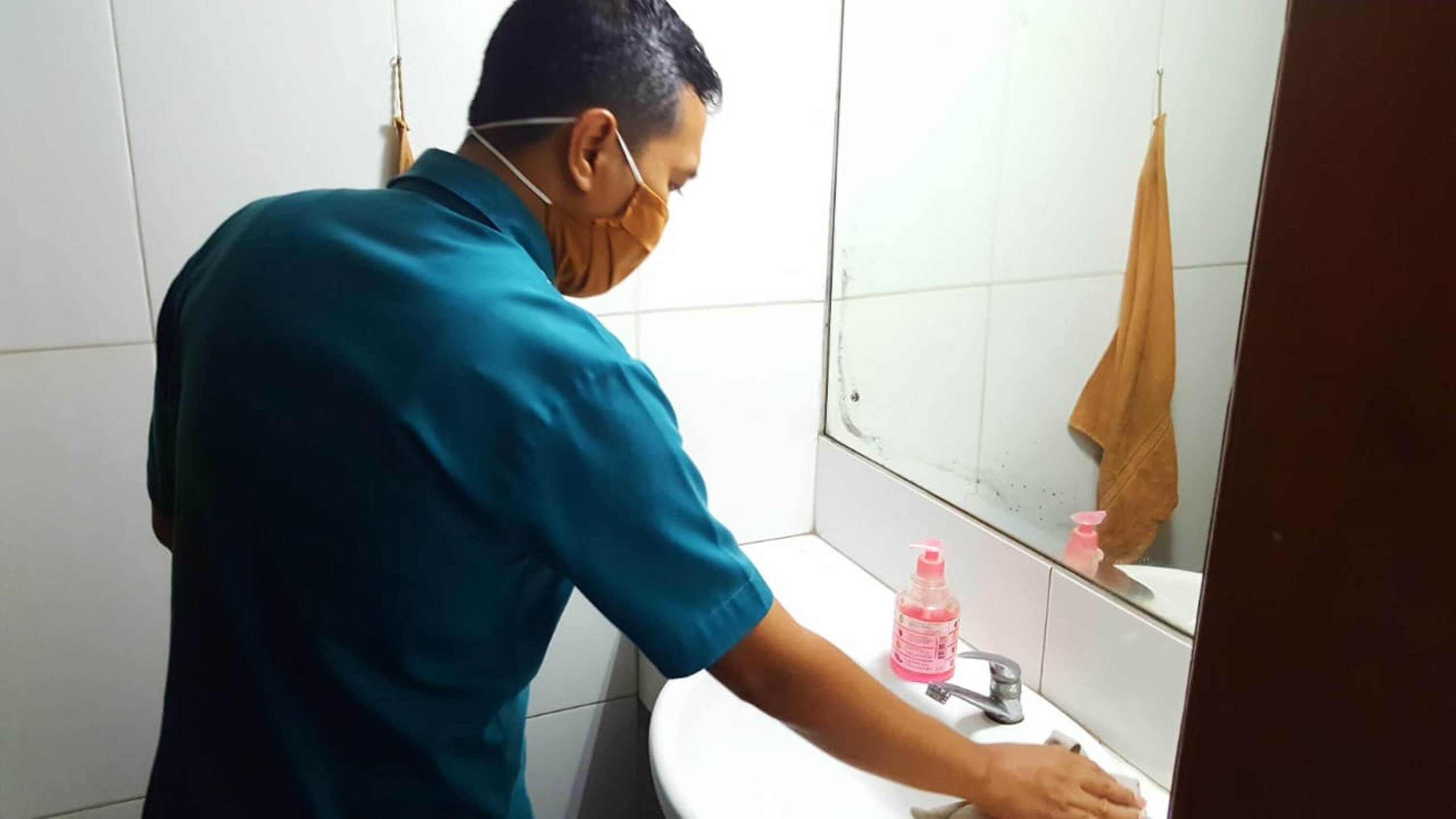 A person washing hands at a sink in a bathroom with a mirror, a towel hanging on the wall, and a bottle of soap.
