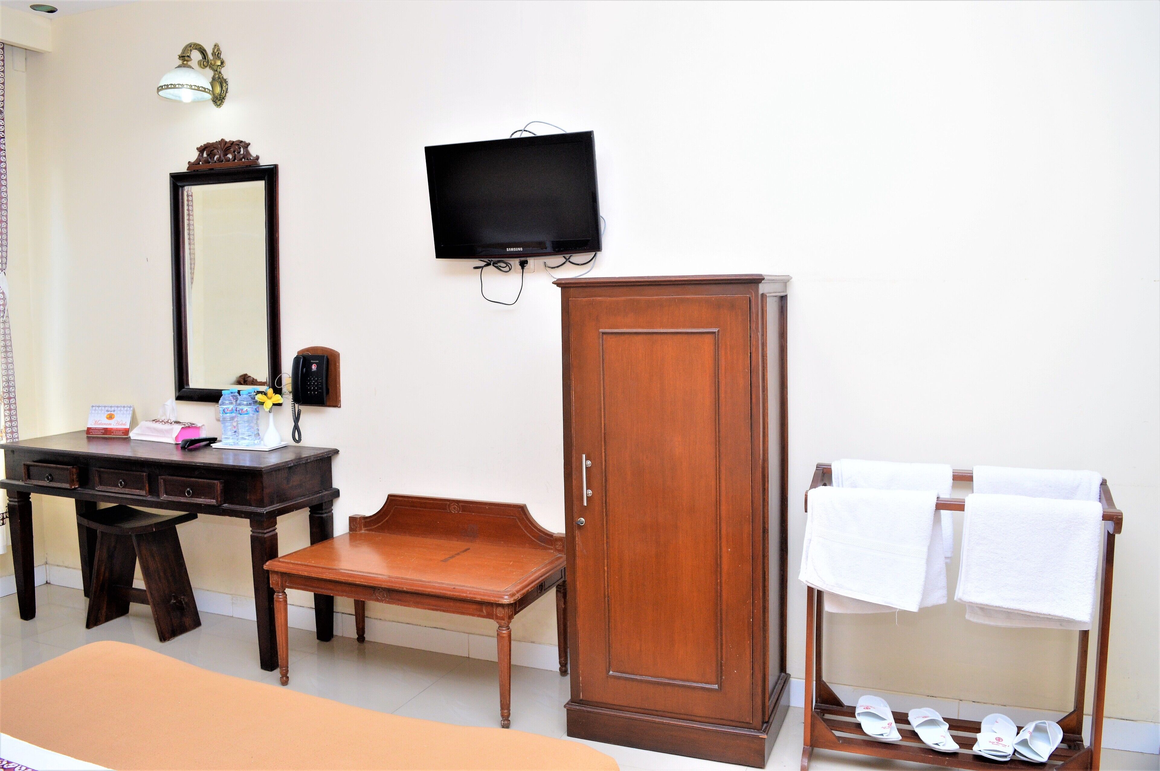 Habitación de hotel con un escritorio de madera, un armario, un televisor montado en la pared y un zapatero con toallas.
