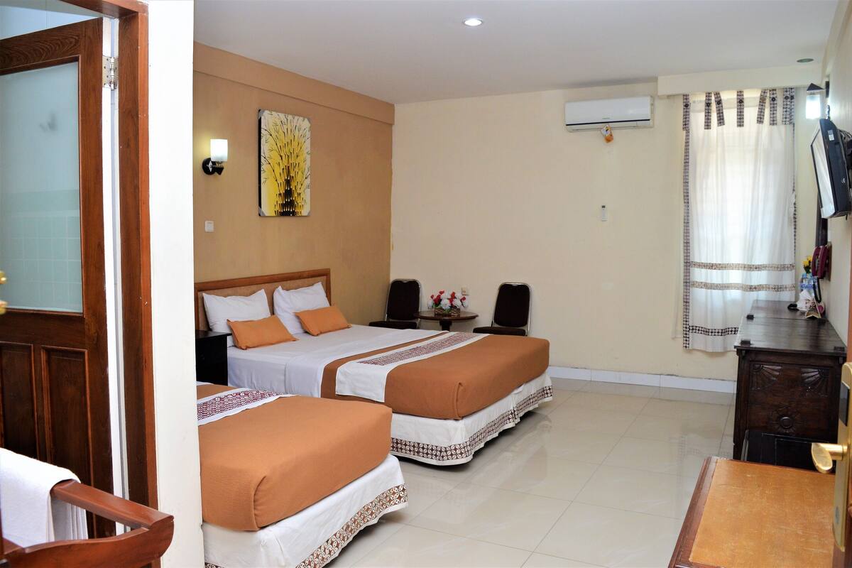 Une chambre d’hôtel avec deux lits, une chaise en bois, une petite table, un climatiseur fixé au mur et un rideau.