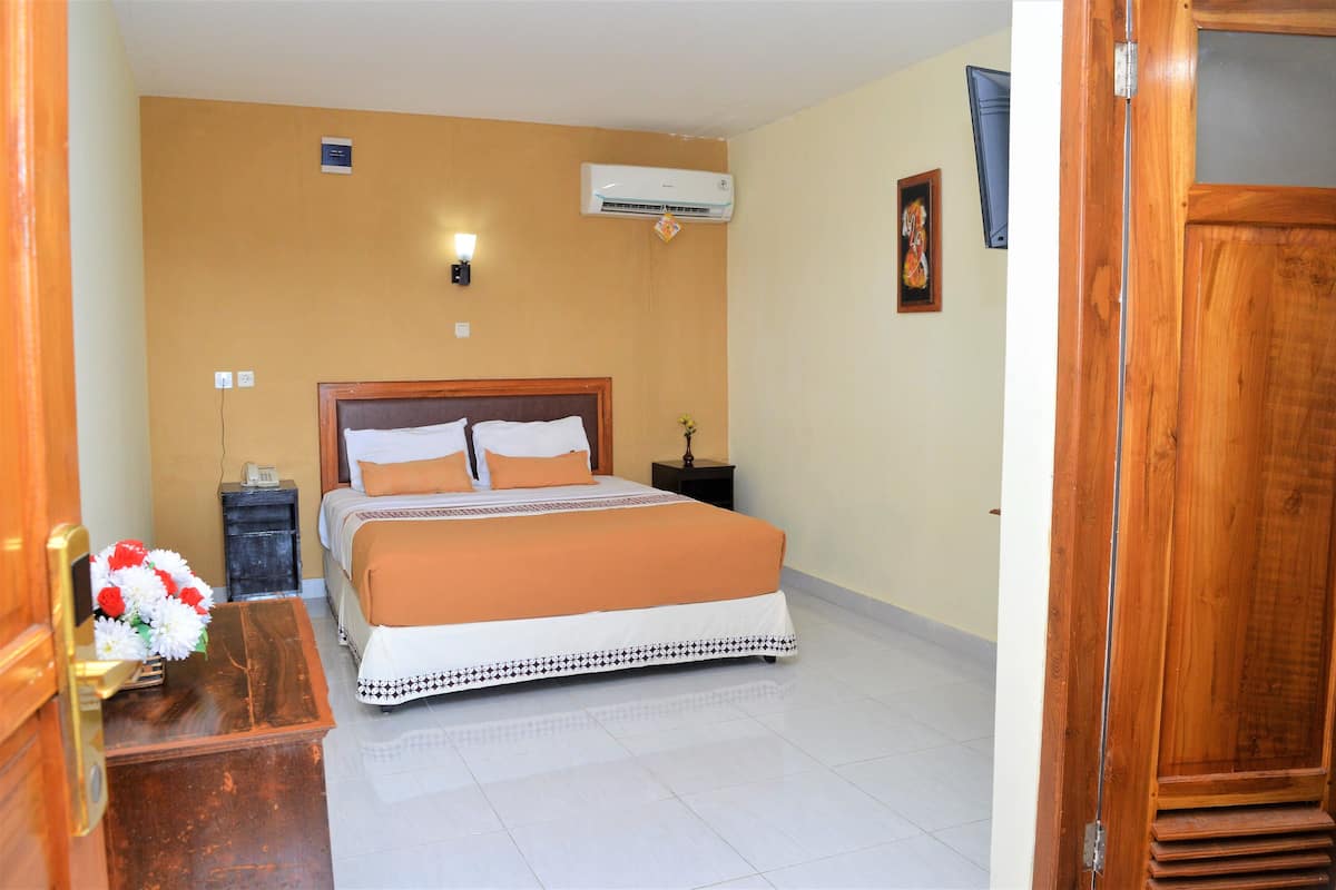 Une chambre d’hôtel avec un lit, une table de chevet, un climatiseur fixé au mur, une porte en bois et une petite table sur laquelle est posé un vase de fleurs.