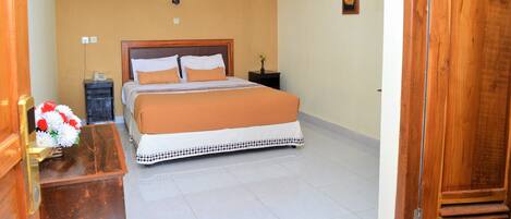A hotel room with a bed, a nightstand, a wall-mounted air conditioner, a wooden door, and a small table with a vase of flowers.