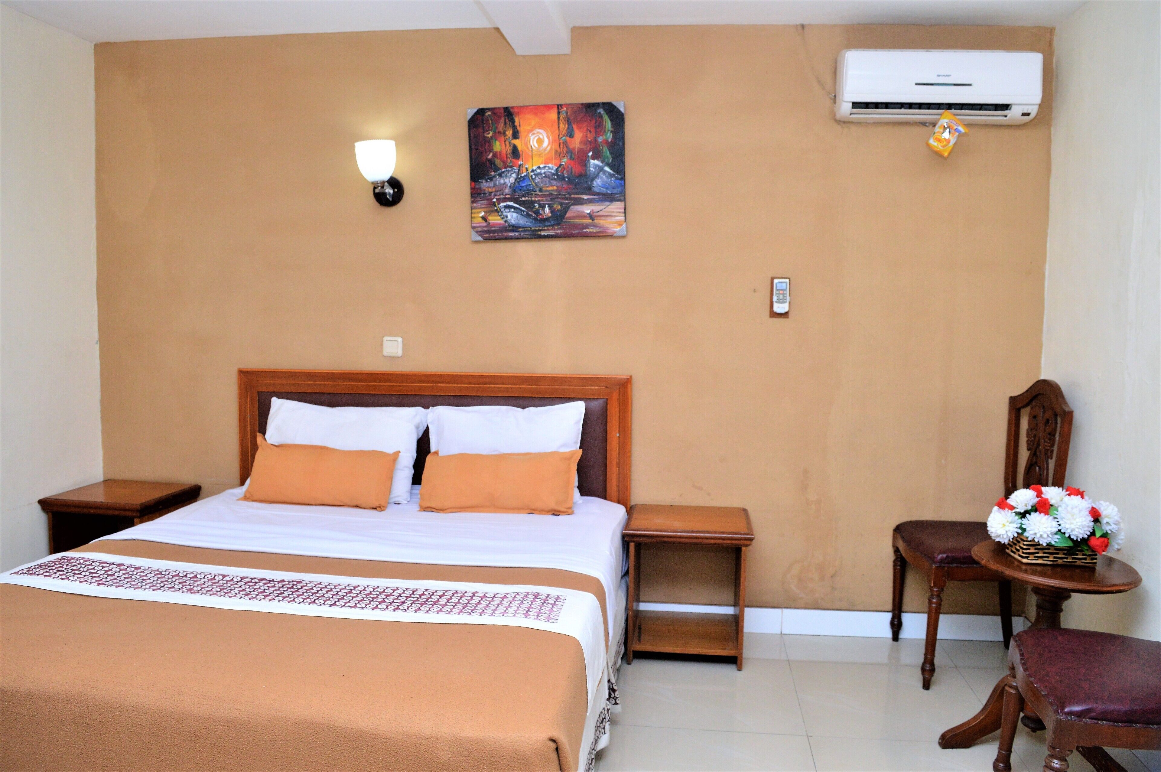 A hotel room with a bed, a nightstand, a chair, a small table with flowers, an air conditioner, and a painting on the wall.