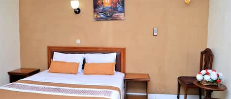 A hotel room with a bed, a nightstand, a chair, a small table with flowers, an air conditioner, and a painting on the wall.