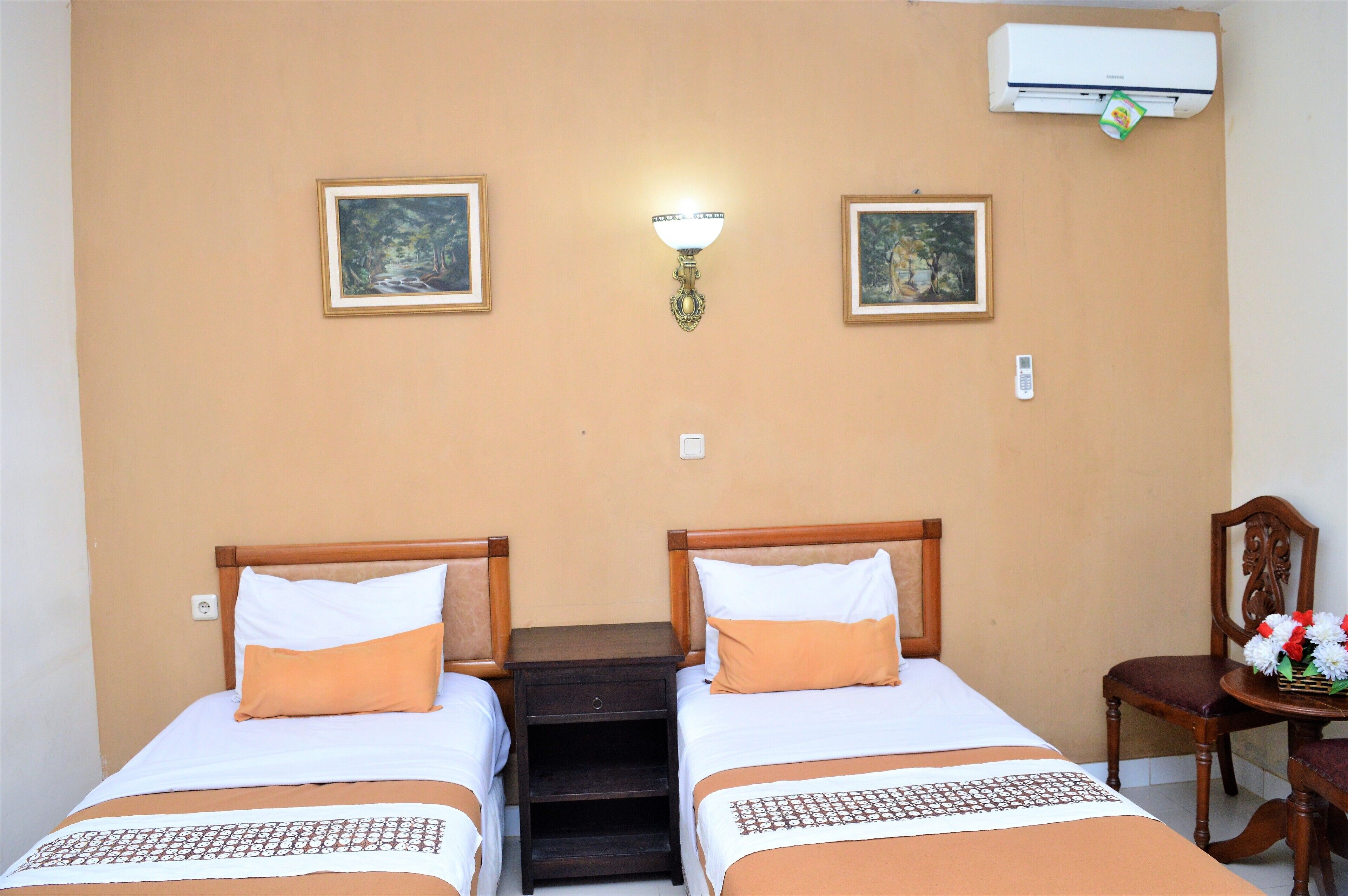 Habitación de hotel con dos camas, una mesita de noche, una lámpara de pared y una silla con un arreglo floral.