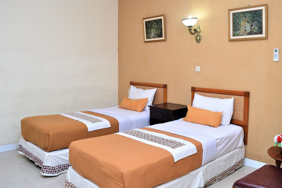 Une chambre d’hôtel avec deux lits, chacun recouvert d’une couverture marron et agrémenté d’oreillers blancs. Deux appliques murales et deux tableaux encadrés ornent le mur.