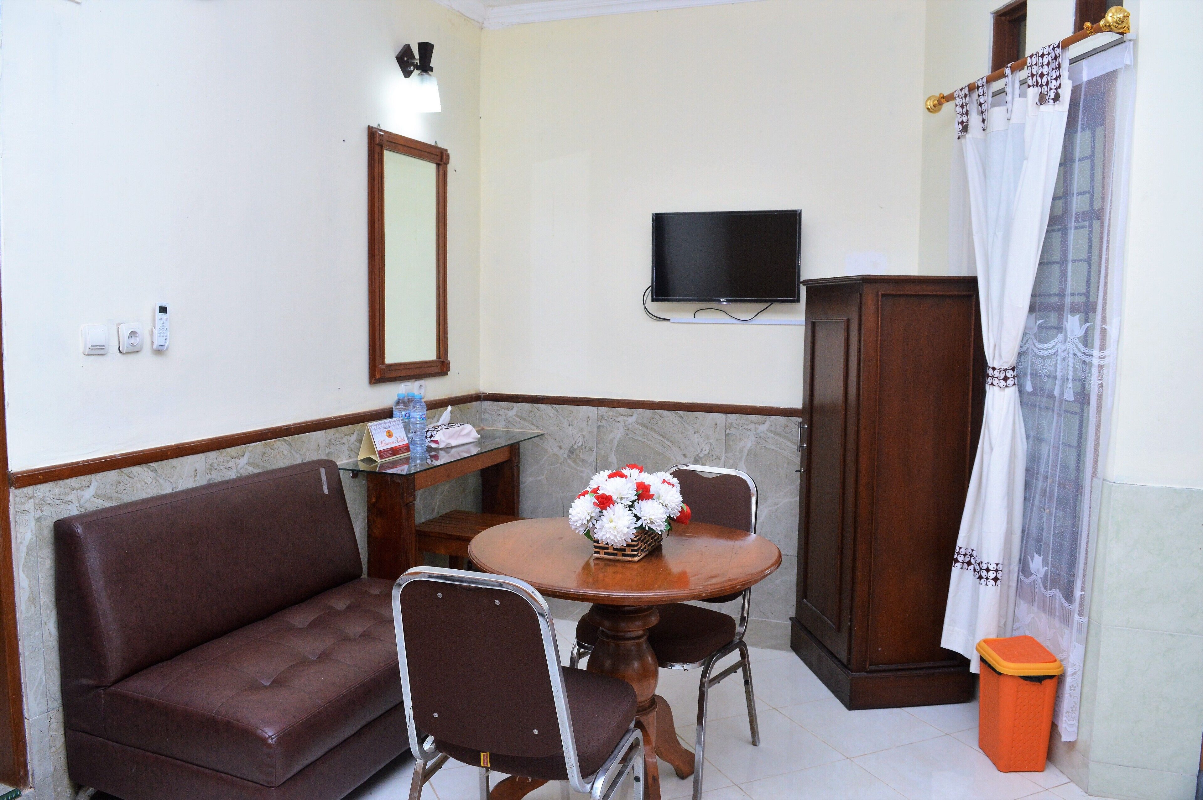 A room with a brown sofa, a wooden table with chairs, a TV mounted on the wall, and a small trash bin.