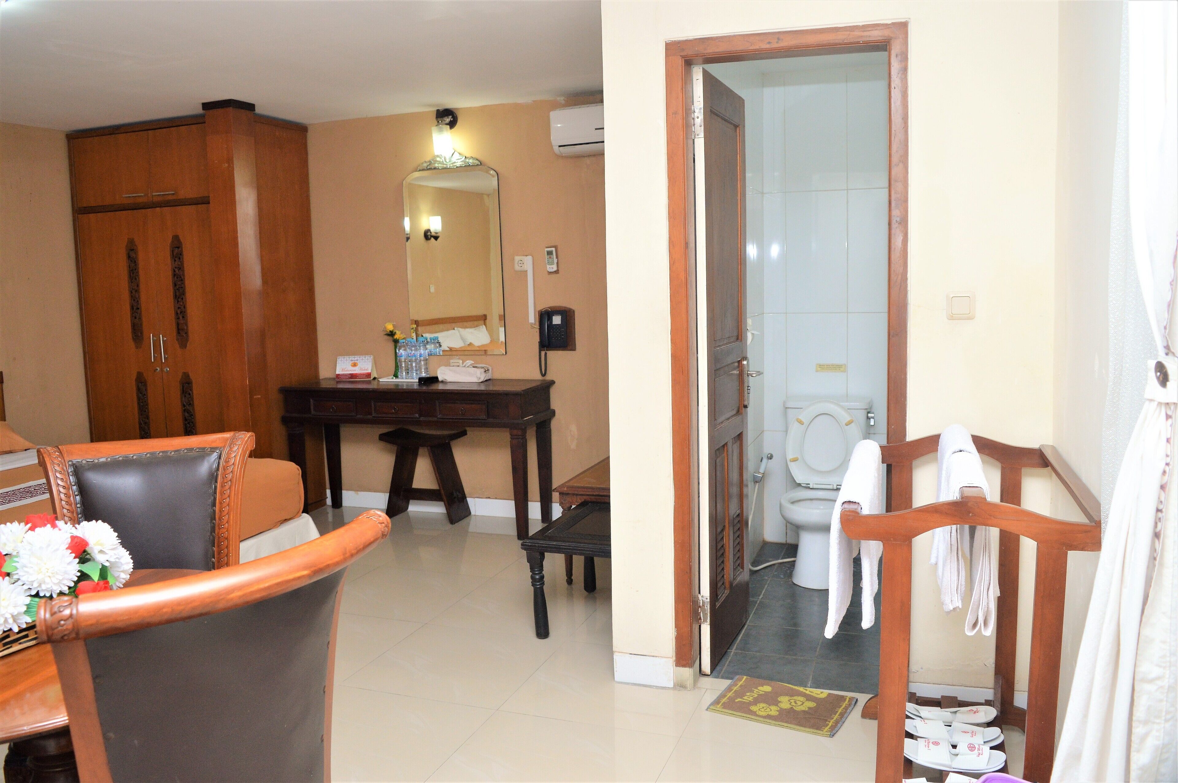 A hotel room with a wooden wardrobe, a desk with a mirror, a telephone, and a bathroom with a toilet and towel rack.