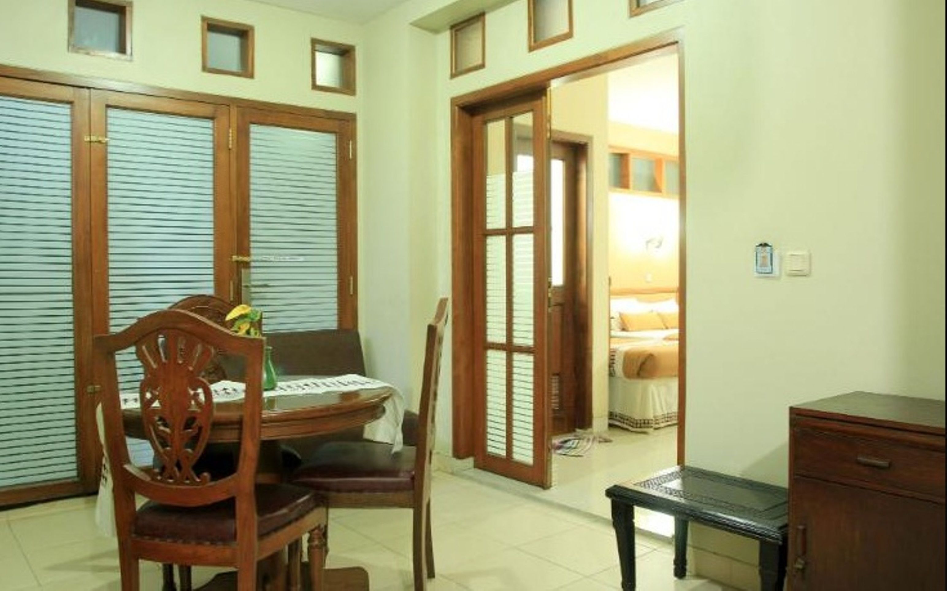 A room with a wooden dining table, chairs, a small cabinet, and a bed in the adjacent room.