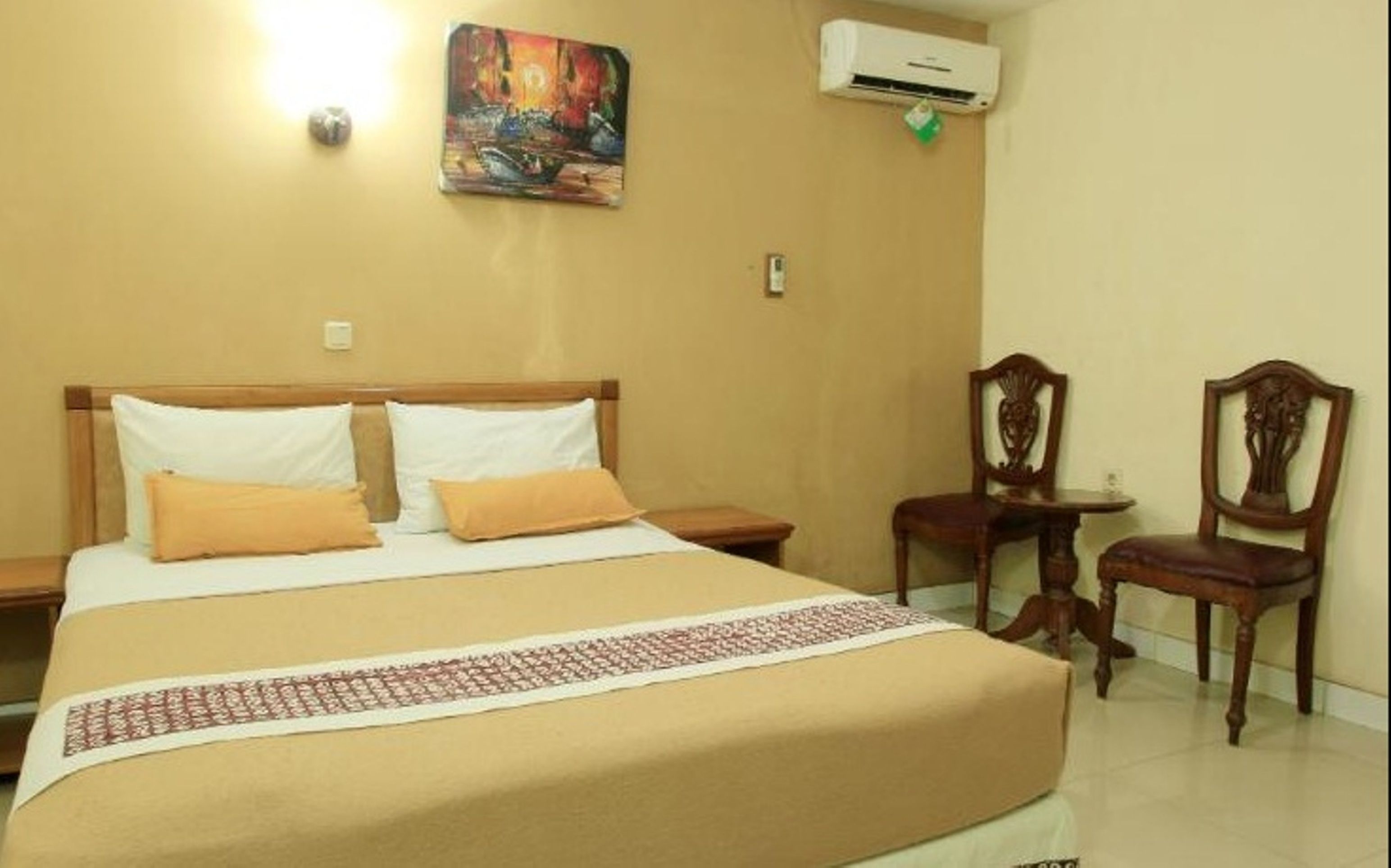 A hotel room with a bed, two chairs, a small table, an air conditioner, and a painting on the wall.