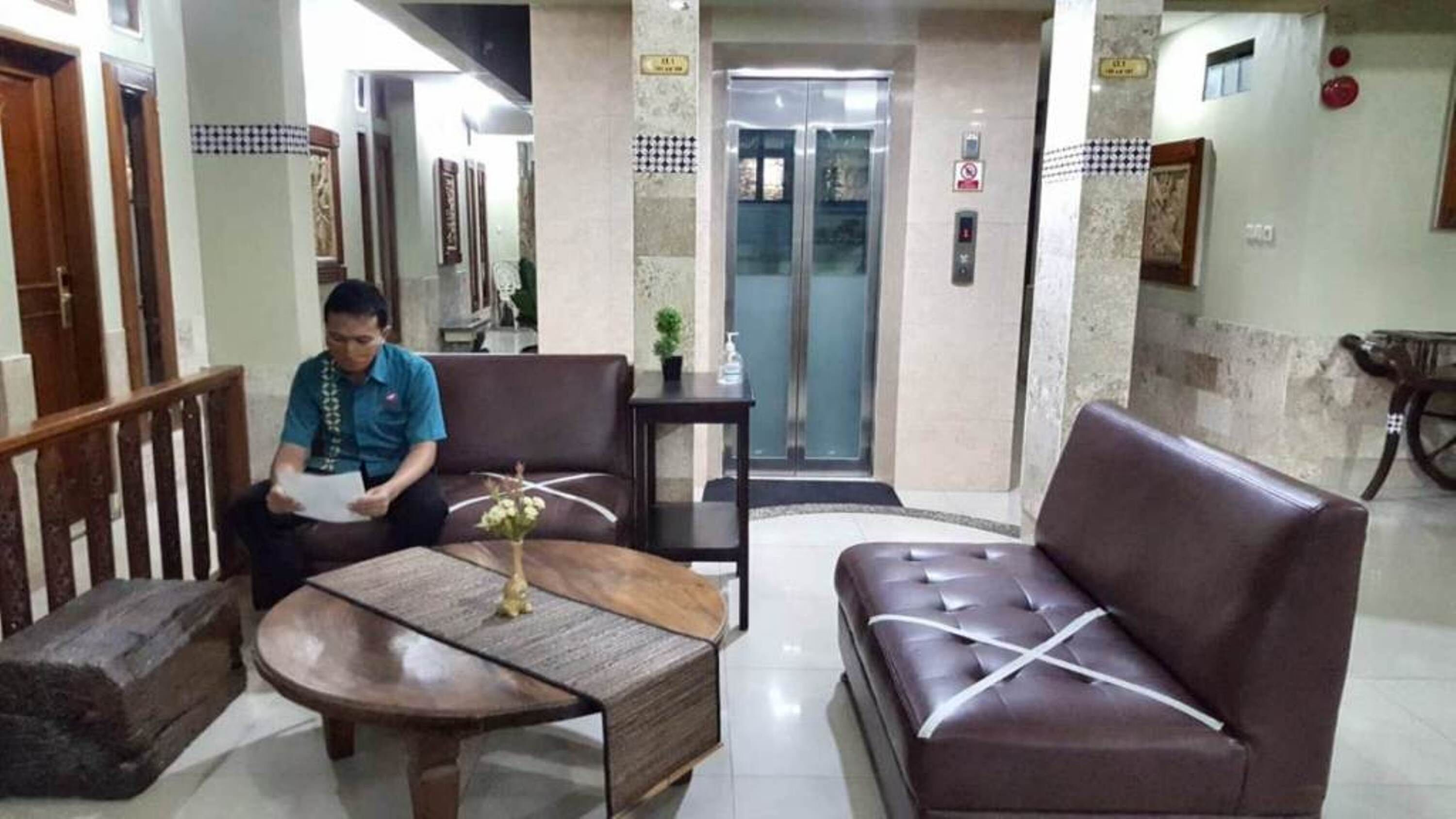 A reception area with a brown leather sofa, a wooden coffee table, and a small potted plant. There is a reception desk with a person seated behind it, and an elevator in the background.