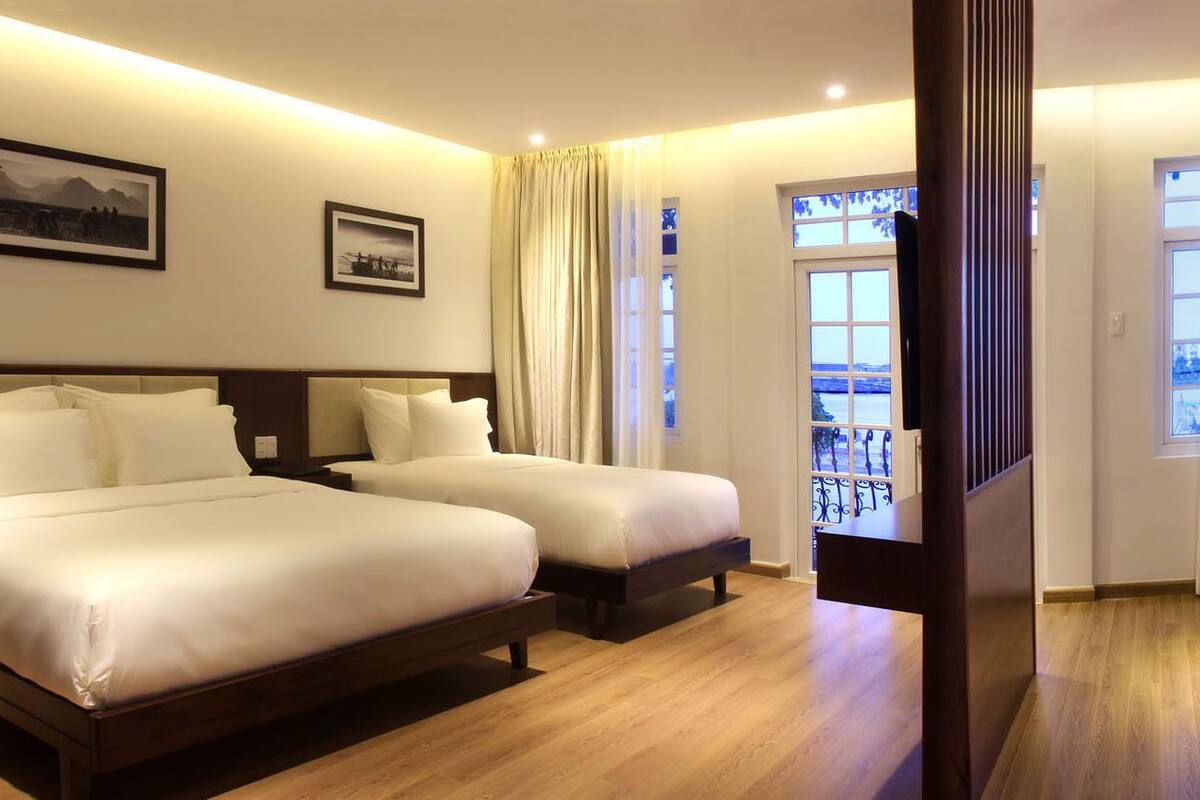 Quarto de hotel com duas camas, piso de madeira, uma janela grande com vista e duas imagens emolduradas na parede.