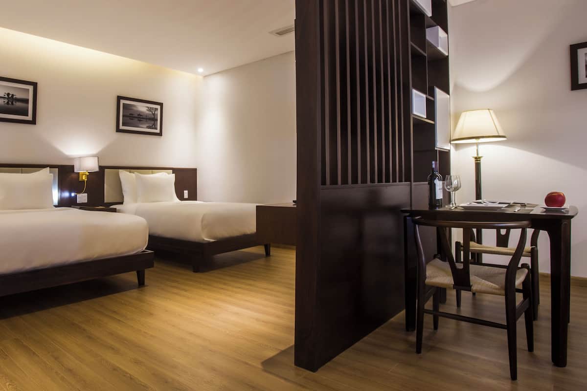 Quarto de hotel com duas camas, piso de madeira, uma escrivaninha com cadeira e uma luminária.