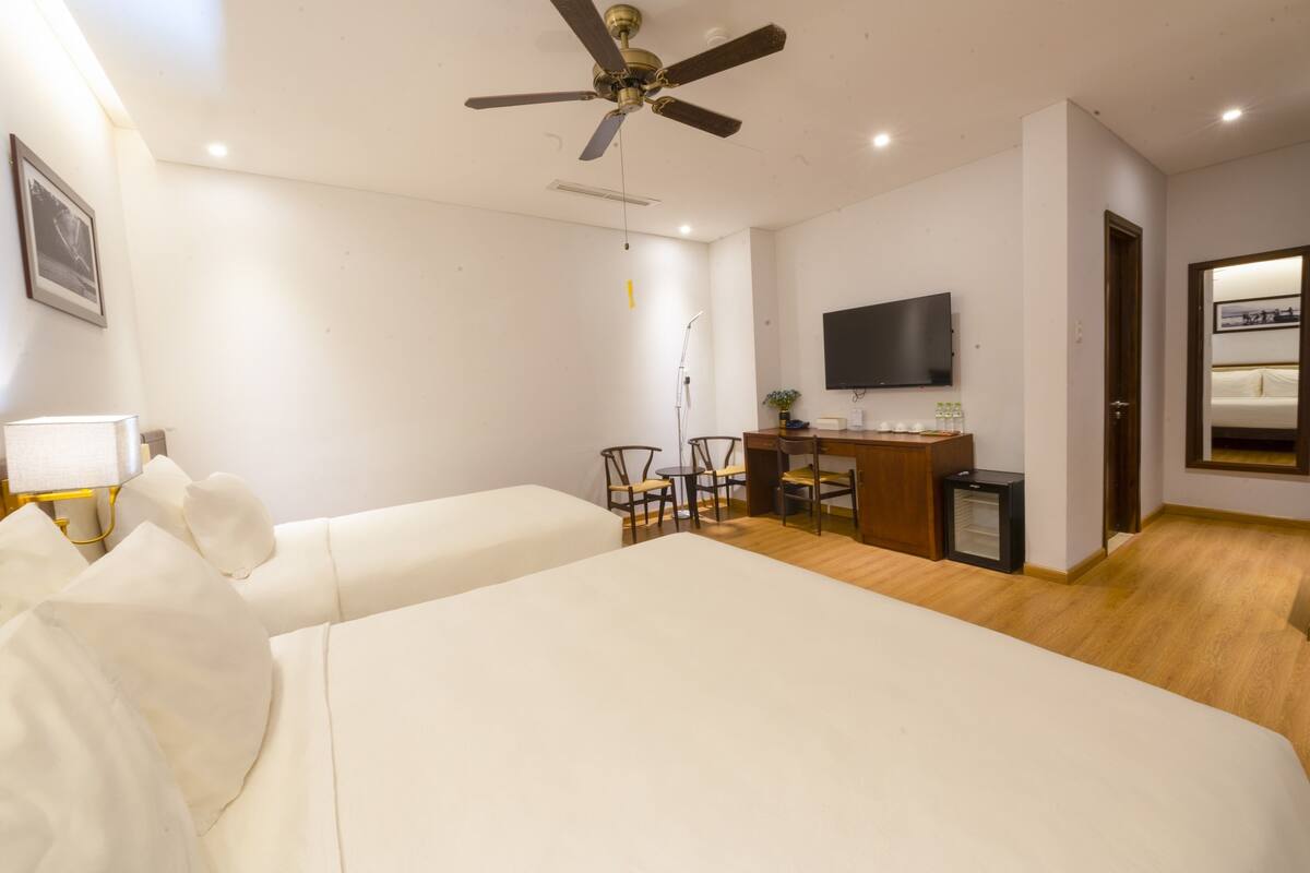 Quarto de hotel com duas camas, uma escrivaninha, uma cadeira, uma televisão e um ventilador de teto.