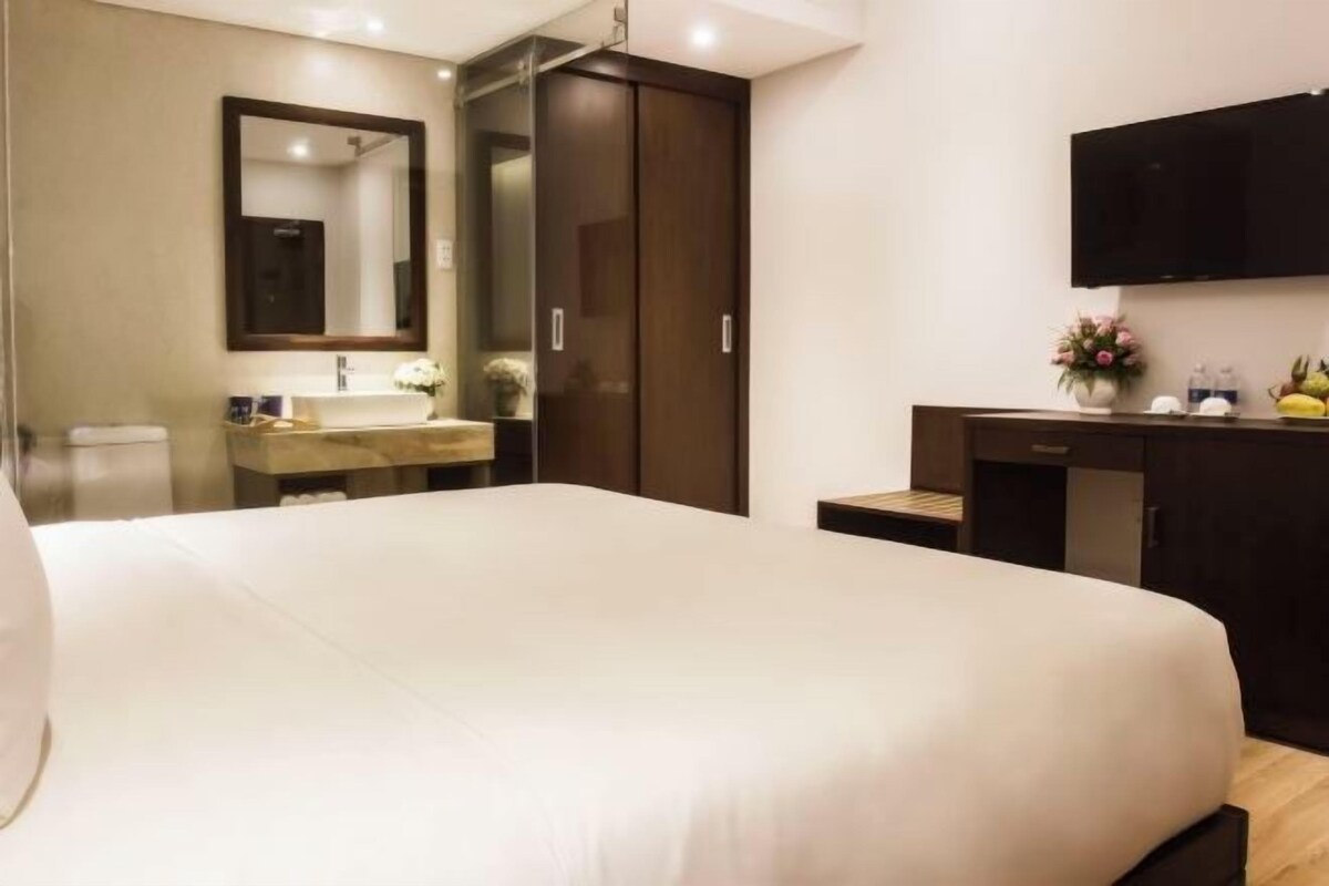A hotel room with a large bed, a bathroom with a sink and mirror, a wardrobe, and a flat-screen TV mounted on the wall.