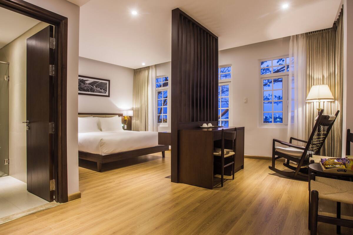 Quarto de hotel com cama, escrivaninha, cadeira e janela com cortinas.