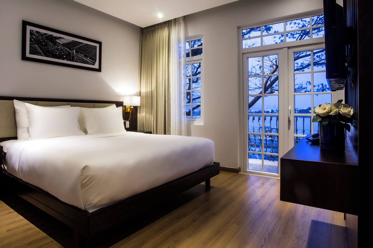 Uma cama bem arrumada em quarto de hotel, com uma janela grande, um quadro na parede e um vaso com flores na mesa de cabeceira.