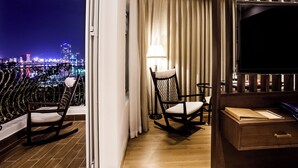 A balcony with a city view, a rocking chair, a chair with a lamp, and a TV. - Di lusso Boutique Hotel Riverside Danang (Da Nang)