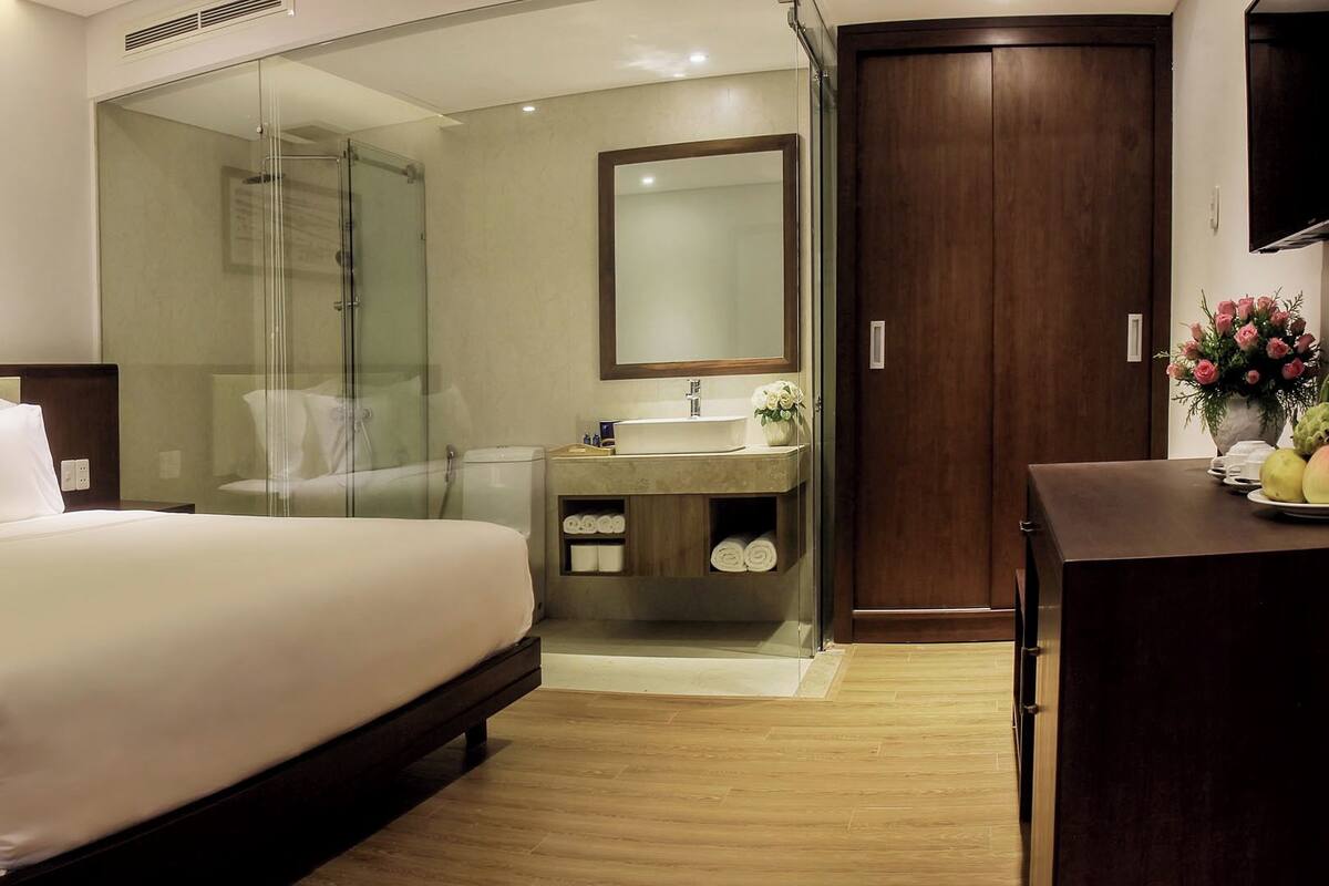 Quarto de hotel com cama, chuveiro em box de vidro, penteadeira com espelho e cômoda de madeira.