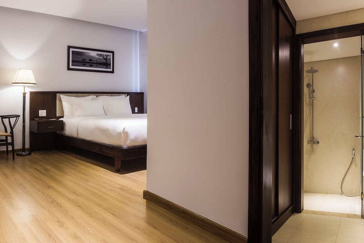 Quarto de hotel com cama, mesa de cabeceira, luminária, cadeira e área de chuveiro.