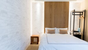 A modern bedroom with a white brick wall, a wooden headboard, a neatly made bed with white and beige bedding, a small wooden nightstand, and a nightstand with a blue kettle. - Tianli Hotel (Tainan)