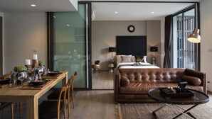 A modern hotel room with a dining area, a leather sofa, a bed with a large TV, and a view through a glass door.