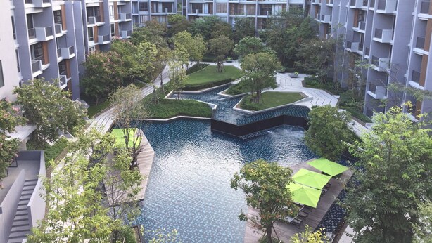 A modern residential complex with multiple high-rise buildings, a central landscaped area with a water feature, and outdoor seating.