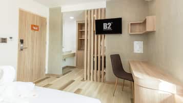 A hotel room with a wooden door, a TV on the wall, a desk, a chair, and a bed.