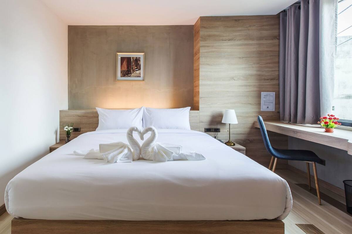 A hotel room with a neatly made bed, a desk with a chair, a nightstand, and a wall-mounted lamp.