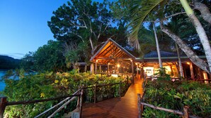 A wooden pathway leading to a building with a thatched roof, surrounded by lush greenery and palm trees. - Savasi Island Resort (Savusavu)